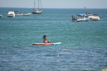 Person paddling on a colorful paddleboard among boats in a sunny coastal location near clear blue water
