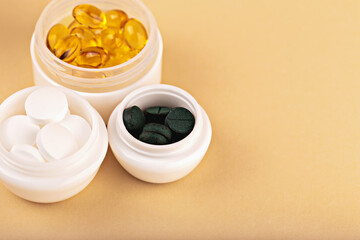 Three white containers filled with yellow capsules, white tablets and green spirulina pills on beige background. Concept of daily vitamins and nutrition care. Supplement tablets in white containers