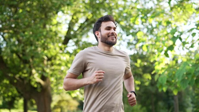 Handsome male runner jogging in an urban city park and looking smart watch. Young athlete using fitness tracker. Sportsman checking result. Adult fit man in T-shirt enjoys running outdoors in nature
