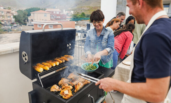 Happy friends cooking corncobs and chicken at barbecue rooftop dinner - Multiracial people having fun during summer time eating and laughing together - Main focus on woman face