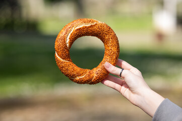 A hand holds a sesame simit outdoors in front of a park on a sunny day.