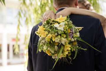 Braut umarmt Bräutigam mit Hochzeitsstrauß aus gelben Blumen im Sommerlicht