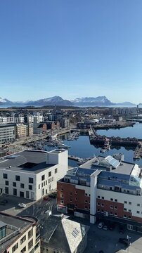 Panoramic view of Bodo, Norway, on a sunny spring day. Snow-covered mountains in the background contrast with the clear streets, harbor, and modern city below.