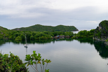 Fototapeta premium Calm bay with sailboat anchored near a village surrounded by forested hills