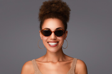 Stylish African American woman with a curly high bun and hoop earrings, smiling in oval-shapped tortoiseshell sunglasses and glittery golden top. Beauty eye wear fashion portrait on a grey background