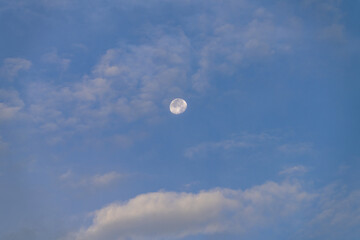 Full moon visible through soft clouds in blue sky during early evening light