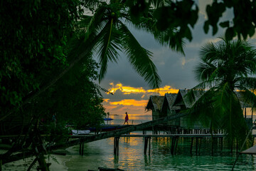 Fototapeta premium Man walking on wooden bridge above calm tropical water with glowing sunset in background