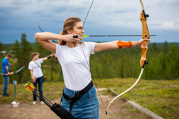 Young woman is practicing archery outdoors, aiming a bow with arrows in a quiver.