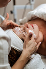 Professional cosmetician applying beauty mask on female client's face. Middle aged woman receiving skincare treatment in beauty spa or salon.