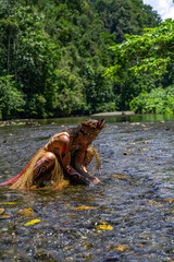 Tribal woman in jungle river kneeling in prayer-like pose, wearing colorful ceremonial body paint