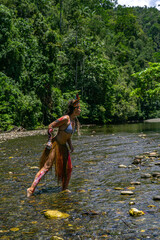 Indigenous woman in traditional feathered headdress and skirt stands confidently in river...