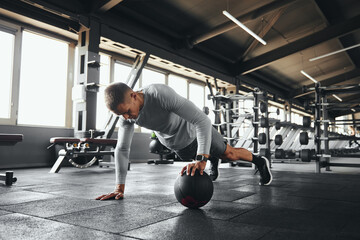 Muscular man doing push up exercise with one hand on medicine ball during intense fitness workout in modern gym