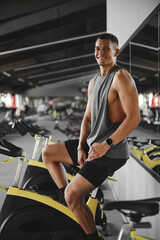 Vertical photo of young man in sportive outfit smiling while sitting on cycling machine in spacious modern gym.