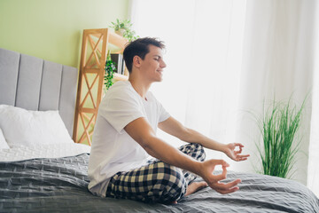 Young man practicing meditation on his bed in a bright and comfortable bedroom for relaxation and...