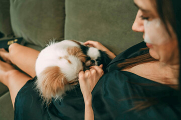 woman in black silk robe relaxing with dogs, smiling while applying under-eye patches, home comfort, concept of skincare and pet bonding