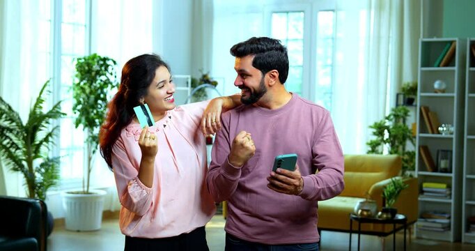 Indian Asian young couple celebrating success while standing in modern living room showing joyful expressions while holding credit or debit card smartphone after online shopping or earning rewards