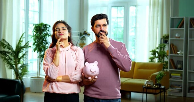 Indian Asian young couple holding a piggy bank, appearing confused and thinking seriously about where to invest or how to utilize their savings smartly and wisely, standing together in living room