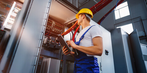 Young electrician works with a tablet standing near an industrial fuse board cabinet. Young...