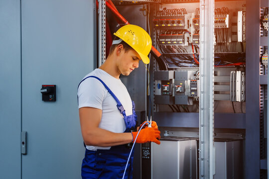 Young electrician works with pliers standing near an industrial fuse board cabinet. Installation of electrical wiring at the factory.