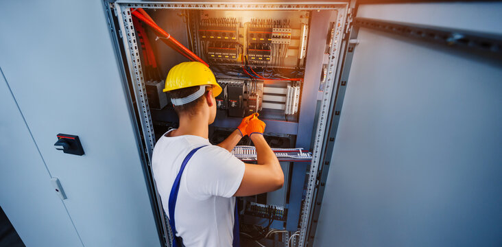Electrician with screwdriver tighten up switching electric actuator equipment in fuse box. Young electrician is repairing installation wiring in switchboard booth at an enterprise. View from the back.