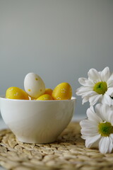  Bright and cheerful Easter-themed still life with a white ceramic bowl of yellow and white decorative eggs and fresh white daisies on a woven placemat. 