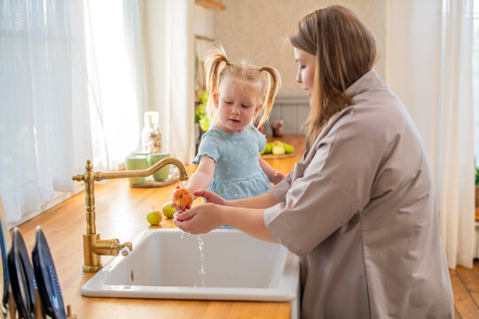 Parent and child washing a ripe apple under the kitchen sink faucet. The child, wearing a blue dress, seems engaged and curious as they hold the fruit amid a cozy kitchen setting