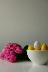 Minimalist spring composition with yellow polka dot Easter eggs in a white ceramic bowl next to a bouquet of pink roses. Clean background and soft light make this image perfect for Easter 