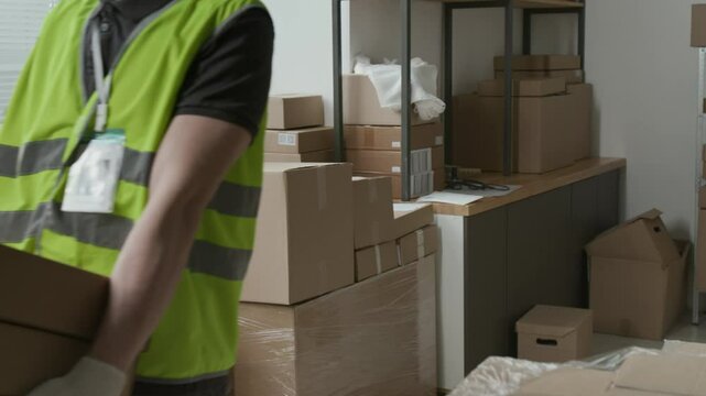 Caucasian male employee wearing jeans and safety vest over t-shirt carrying three cardboard boxes during workday in storeroom