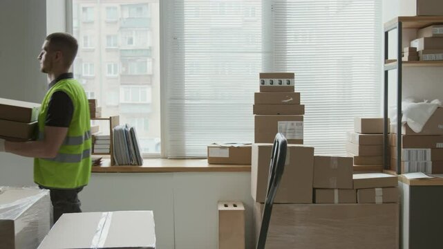 Side view of young man wearing green high-visibility vest and work gloves slowly walking across storeroom carrying three boxes