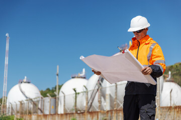 A construction engineer in protective gear examines blueprints at a gas storage facility. He holds...