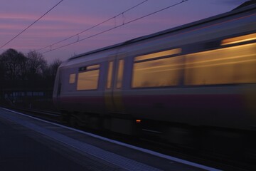 Train speeds past platform, illuminated windows reflecting twili