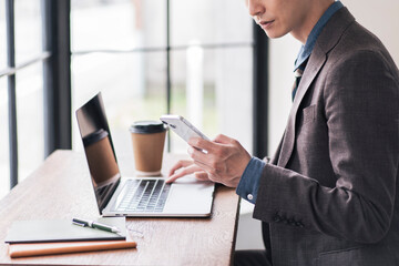 ノートパソコンを開き、スマートフォンを見ながら作業をしている日本人男性。