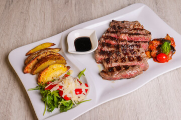 Ribeye di manzo cotta al sangue, scaloppata e servita in una braceria con contorno di patate al forno, rucola, scaglie di formaggio, pomodori e salsa bbq
