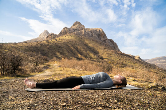 Young sporty woman practicing yoga, lying with eyes closed in Dead Body or Corpse pose, Savasana exercise. Morning fitness workout in nature, resting after yoga in sunny day in mountains, wellness