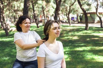 Fototapeta premium Smiling mother tying teenage daughters hair while sitting outdoors