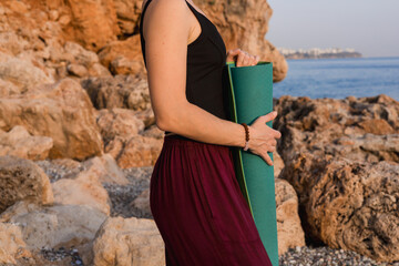 Young caucasian woman holding rolled up green yoga mat against background of sea rocks.