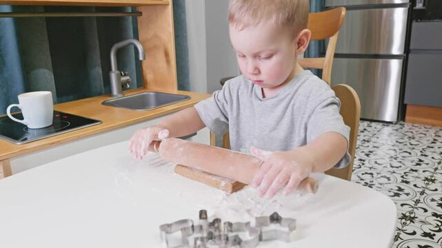A toddler enthusiastically rolls out dough on a floured surface, surrounded by cookie cutters. The cozy kitchen setup captures the joy of holiday baking and family traditions