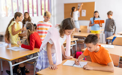 Obraz premium Positive friendly tween girl helping classmate sitting at school desk to complete written assignment in notebook during break before lesson ..