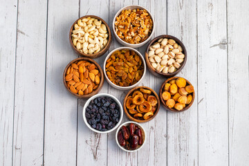 an assortment of dried fruits and nuts arranged in bowls on a white wooden surface, including dates, cashews, almonds, raisins, figs, apricots, and walnuts.