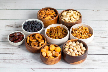 an assortment of dried fruits and nuts arranged in bowls on a white wooden surface, including dates, cashews, almonds, raisins, figs, apricots, and walnuts.