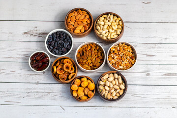 an assortment of dried fruits and nuts arranged in bowls on a white wooden surface, including dates, cashews, almonds, raisins, figs, apricots, and walnuts.