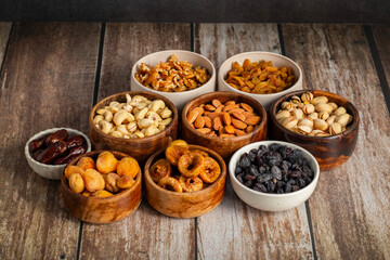 an assortment of dried fruits and nuts arranged in bowls on a white wooden surface, including dates, cashews, almonds, raisins, figs, apricots, and walnuts.