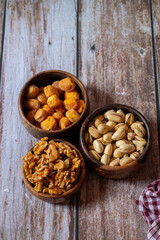 Three wooden bowls filled with dried apricots, pistachios, and walnuts rest on a rustic wooden table beside a red and white checkered cloth.