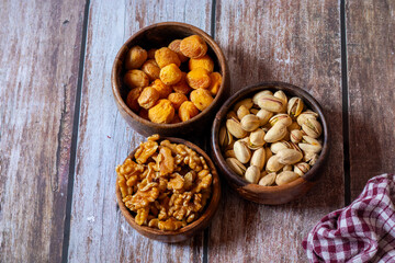 Three wooden bowls filled with dried apricots, pistachios, and walnuts rest on a rustic wooden table beside a red and white checkered cloth.