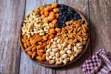 A round wooden plate holds cashews, almonds, pistachios, dried apricots, raisins, and walnuts, neatly arranged on a rustic wooden table beside a red checkered cloth.