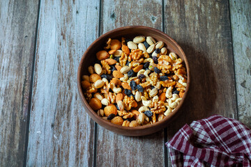 A round wooden plate holds cashews, almonds, pistachios, dried apricots, raisins, and walnuts, neatly arranged on a rustic wooden table beside a red checkered cloth.