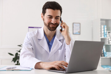 Professional doctor talking on smartphone while working with laptop at white desk in hospital