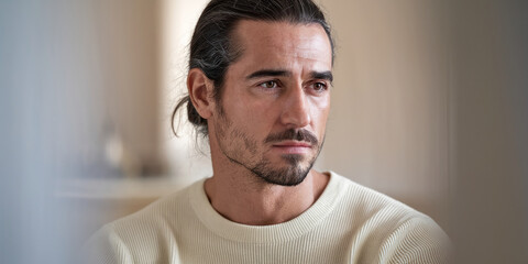 Close-up of a man with brown eyes and long hair pulled back, wearing a cream-colored sweater, conveying thoughtfulness and introspection