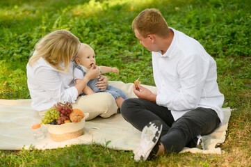 Fototapeta premium Mom, dad and baby enjoy food, picnic in park, eat fruits, feed microdoses to baby. Family leisure, summer vacation and picnic concept