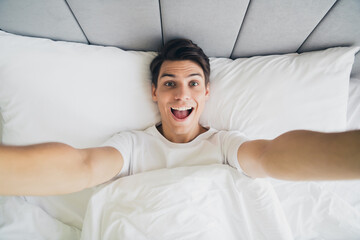 Happy young man in bed wakes up with a smile and outstretched arms in the morning light at a cozy...